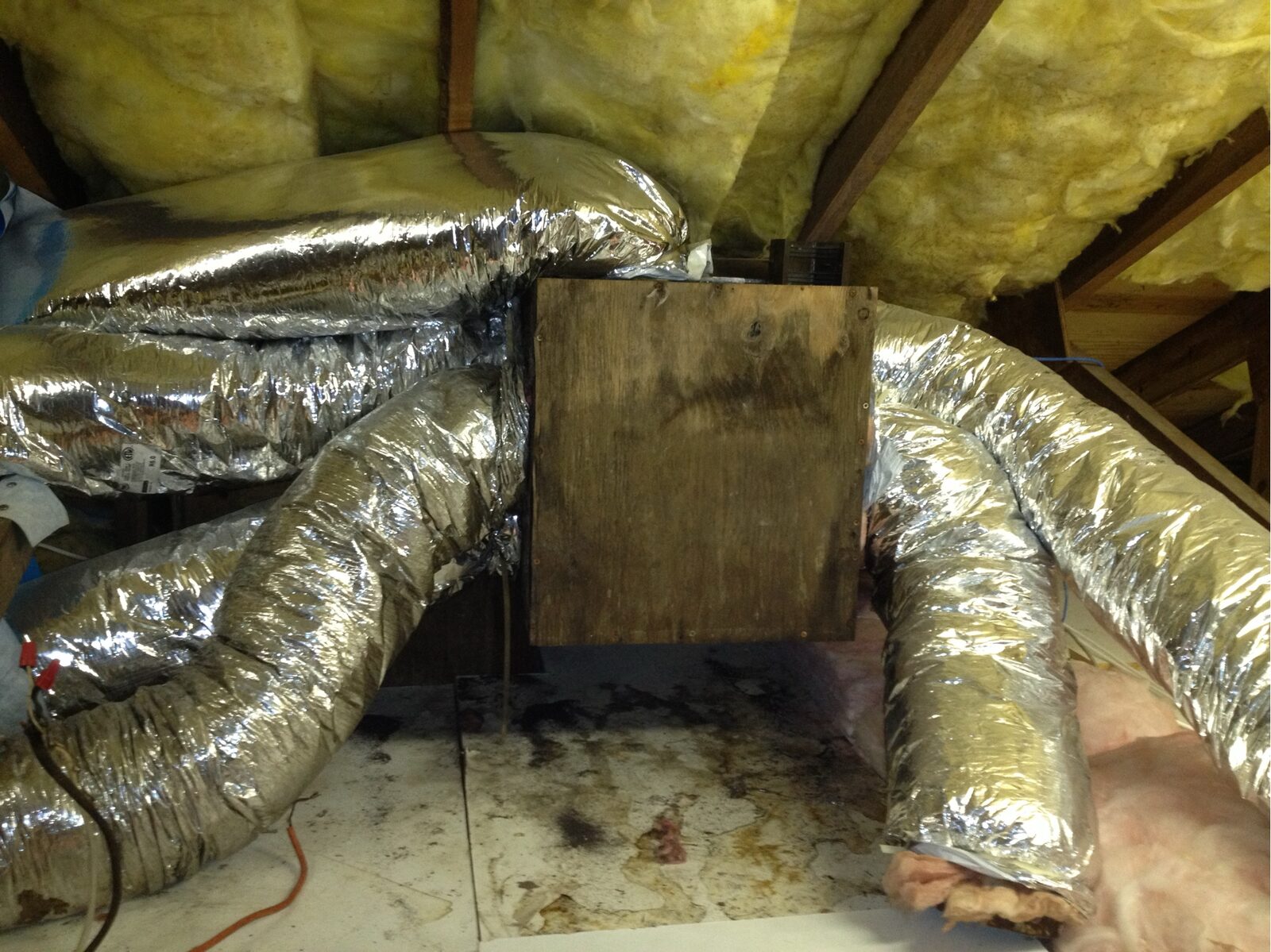 Flexible ductwork and reflective insulation running across rough attic framing above a stained subfloor