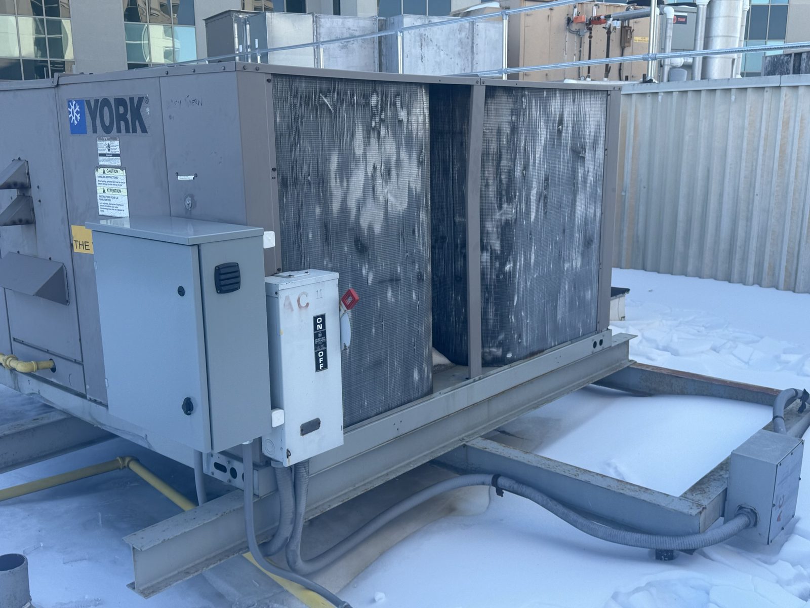 Close-up of the York unit's weathered condenser coils and electrical disconnect, showing age and wear on a snowy rooftop